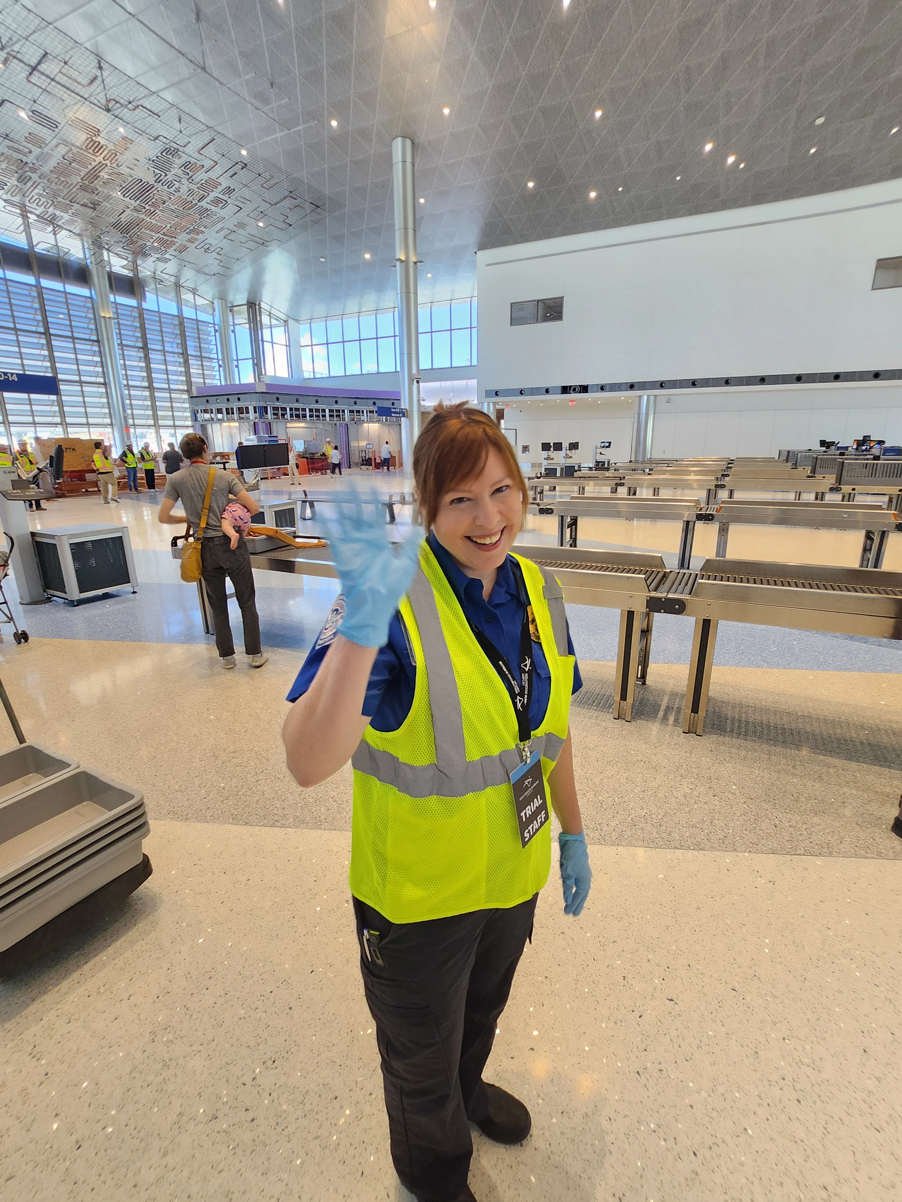 A TSA agent in a yellow safety vest waving and smiling near the security screening lanes